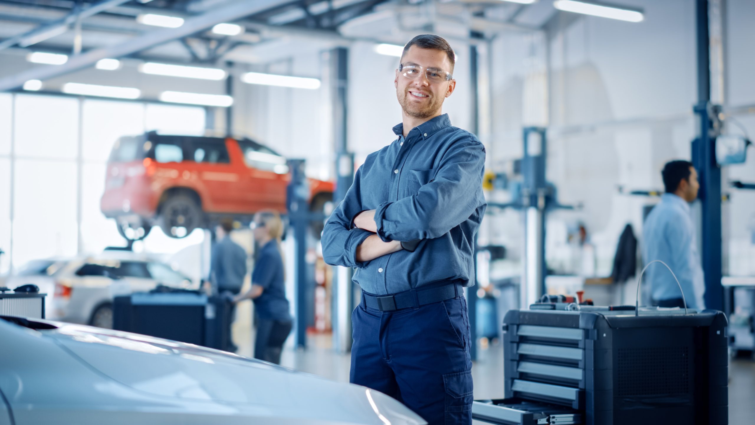 Smiling auto technician wearing safety glasses stands with arms crossed inside a modern auto repair shop, with vehicles on lifts and other mechanics working in the background.