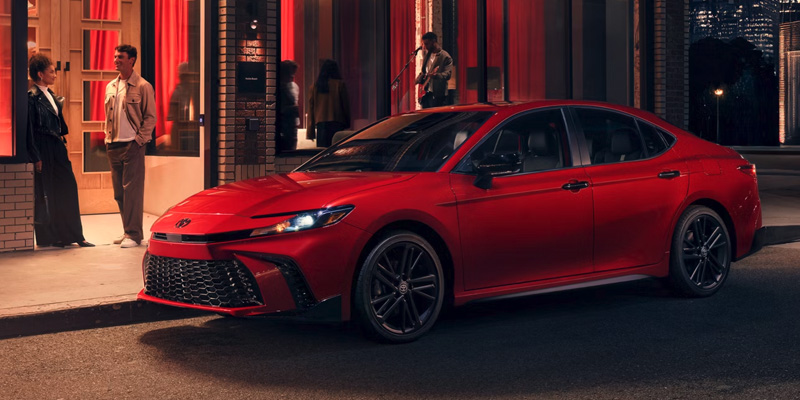 A red Toyota Camry parked on a city street at night, near a couple talking by a building.